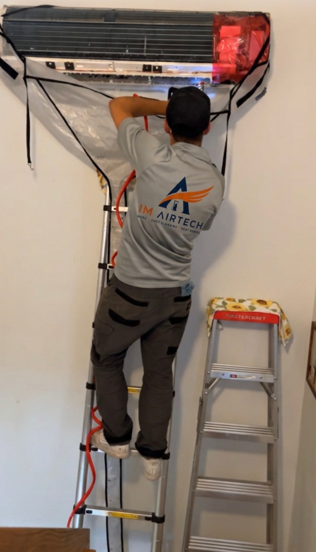ImAirtTech technician on a ladder cleaning a wall-mounted heat pump with a protective cleaning bag installed